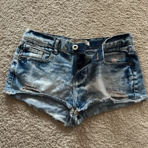 Blå jeansshorts med slitninga - Snygga blå jeansshorts med slitna detaljer och fransig kant. Perfekta för en avslappnad stil. De har en knapp och dragkedja framtill samt fickor både fram och bak.