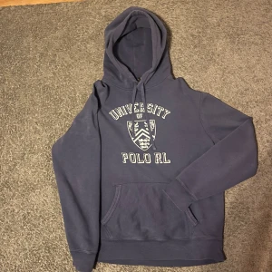 Mörkblå hoodie från Ralph Lauren - Säljer en snygg mörkblå hoodie från Ralph Lauren med texten 'University Polo RL' och ett stilrent emblem på bröstet. Tröjan har en klassisk känguruficka och justerbar huva med dragsko. Perfekt för en avslappnad stil. Nu pris runt 3000kr