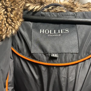 Svart vinterjacka med päls från Hollies - Säljer en snygg svart vinterjacka från Hollies med en lyxig pälsdetalj på huvan. Jackan har dragkedja och praktiska fickor med dragkedjor. Perfekt för kalla dagar när du vill hålla dig varm och stilren. 🖤