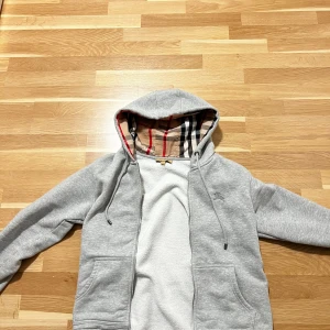 Grå hoodie från Burberry - Säljer en stilren grå hoodie från Burberry med klassiskt rutigt mönster i huvan. Den funkar till både M och L Jackan har dragkedja och broderad logga på bröstet. Perfekt för en avslappnad look. 