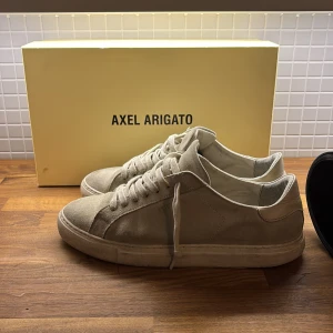Beige sneakers från Axel Arigato - Snygga beige sneakers från Axel Arigato i mocka med klassisk snörning. Perfekta för en stilren look. Skorna har en enkel och tidlös design som passar till många olika outfits. Dem har inga defekter bara att sulan har smuts på sig som enkelt går att ta bort. Hör av dig vid frågor eller funderingar om skorna. Priset kan diskuteras 