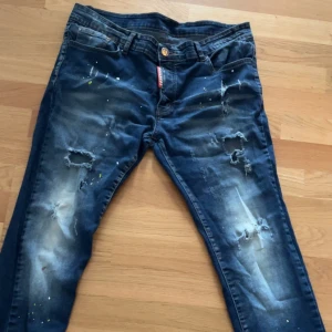 Blå jeans från Dsquared2 - Snygga blå jeans från Dsquared2 med slitna detaljer och färgstänk. De har en knappgylf och klassisk femficksdesign. Perfekta för en trendig look.