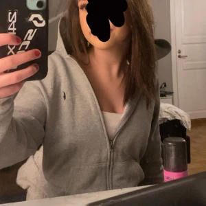 Grå hoodie från Ralph Lauren - Säljer en stilren grå hoodie från Ralph Lauren med dragkedja framtill. Den har en liten broderad logga på bröstet och långa ärmar. Perfekt för en avslappnad look. 