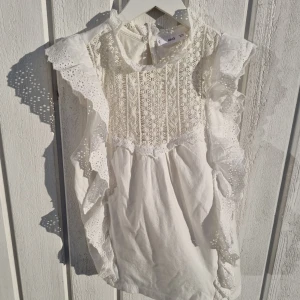 Spetsblus Magno - Säljer en vacker vit blus från Mango med spetsdetaljer och volanger längs ärmarna. Blusen har en elegant spetsdesign framtill och en knapp i nacken. Perfekt för en stilren look.