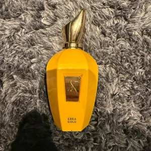 Gul parfymflaska Erea Gold - Säljer en elegant gul parfymflaska från Erea Gold med en unik, skulptural kork i guld. Flaskan har en rektangulär form med en gyllene detalj framtill. Perfekt för den som vill ha en iögonfallande doft på hyllan.