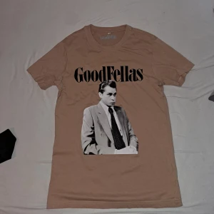 Beige GoodFellas T-shirt - Säljer en beige T-shirt med tryck från GoodFellas. Den har ett svartvitt motiv av en man i kostym och texten 'GoodFellas' i svart. Perfekt för filmälskare som vill ha en stilren look. Den är köpt från foot Locker.