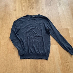 Stickad tröja - Säljer en stilren mörkgrå tröja från Dressmann i mjuk bomull. Tröjan har en klassisk design med långa ärmar och rund halsringning. Perfekt för en avslappnad och bekväm stil.