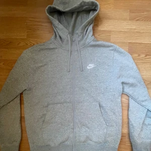 Grå hoodie från Nike - Säljer en stilren grå hoodie från Nike med dragkedja och broderad logga på bröstet. Perfekt för en avslappnad look. Den har en justerbar huva och praktiska fickor framtill. Använd fåtal gånger. Köpt på Stadium för 949kr! Skriv vid funderingar🙌🏼