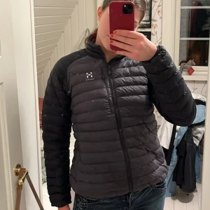 Mörkgrå dunjacka från Haglöfs - Tunn dunjacka som passar bra till våren. Mörkare grå på ärmarna än resten av jackan. Två fickor med dragkedja och lite figursydd passform.