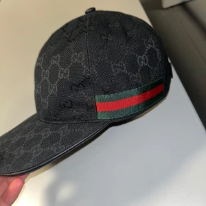 Svart keps med mönster och ränder - Snygg svart keps med diskret mönster och ikoniska ränder i grönt och rött på sidan. Justerbar rem baktill för perfekt passform.