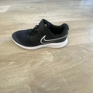 Nike Star Runner 3 Unisex Väg-löparskor - Snygga och bekväma Nike Star Runner 3 unisex löparskor i svart och mörkgrått. Skorna har en vit sula och en ikonisk Nike-logga på sidan. De är utrustade med snörning och en praktisk kardborreband för extra stöd.