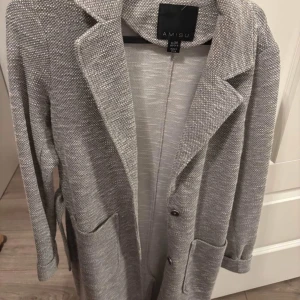 Grå kappa från Amisu - Snygg grå kappa från Amisu i storlek M. Den har en klassisk krage och knappar framtill. Kappan är långärmad och har praktiska fickor samt ett bälte i midjan för en stilren look.