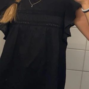 Svart blus med spetsdetaljer - Snygg svart blus med spetsdetaljer och korta ärmar. Perfekt för en stilren look. Blusen har en fin struktur och är lätt att matcha med olika outfits.