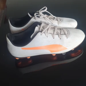 Vita fotbollsskor från Puma 37,5 - Snygga vita fotbollsskor från Puma med orange detaljer och snörning. Skorna har en klassisk design med dobbar för bra grepp på planen. Perfekta för fotbollsträning och matcher.