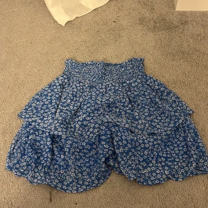 Blå blommig kjol från Pigalle - Söt blå kjol med vita blommor från Pigalle. Kjolen har en volangdetalj och en elastisk midja för en bekväm passform. Perfekt för sommarens alla tillfällen!