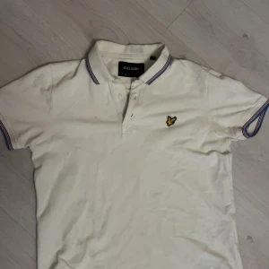 Vit pikétröja från Lyle & Scott - Säljer en klassisk vit pikétröja från Lyle & Scott med korta ärmar och en broderad logotyp på bröstet. Tröjan har en stilren design med färgade ränder på kragen och ärmsluten. Perfekt för en avslappnad stil.