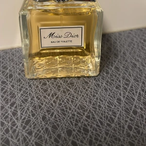 Miss Dior Eau de Toilette - Säljer en elegant flaska Miss Dior Eau de Toilette. Flaskan har en klassisk fyrkantig form med en stilren etikett och en vacker rosett på korken. Doften är fräsch och feminin, perfekt för den som älskar lyxiga parfymer. En tidlös doft som passar alla tillfällen.