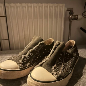 Olivgröna sneakers med nitar - Säljer ett par olivgröna sneakers med coola nitar och slitna detaljer. Skorna har en klassisk vit sula med svarta ränder och snörning framtill. Perfekta för en edgy look!