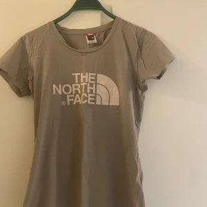 Beige t-shirt från The North Face - Säljer en snygg beige t-shirt från The North Face med deras ikoniska logga tryckt på bröstet och en mindre logga på ryggen. Perfekt för en avslappnad stil. T-shirten är kortärmad och gjord i ett mjukt material.