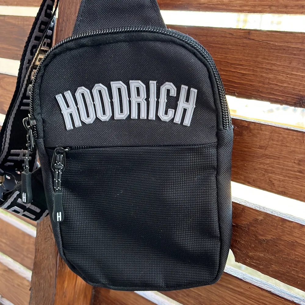 Snygg svart axelväska från Hoodrich med justerbar axelrem och dragkedja. Perfekt för att bära med sig det viktigaste. Väskan har en stilren design med logotypen tydligt tryckt på framsidan och remmen. Aldrig använd.. Laukut & Käsilaukut.