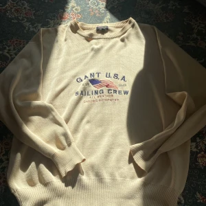 Beige tröja från GANT - Säljer en beige tröja från GANT med tryck av amerikansk flagga och texten 'GANT U.S.A. SAILING CREW'. Tröjan har långa ärmar och ribbade muddar vid ärmslut och nederkant. Perfekt för en avslappnad stil.