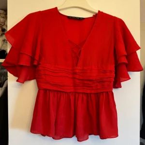 Röd blus från Zara Basic - Säljer en röd blus från Zara Basic med volangärmar och v-ringning. Blusen har en fin detalj med veckad midja och är perfekt för en färgglad look.