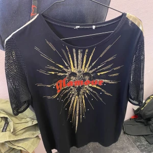 Svart t-shirt med nätärmar och tryck - Snygg svart t-shirt med glittrigt guldtryck och texten 'Glamour' i rött. T-shirten har korta nätärmar och en rund halsringning. Perfekt för en cool och trendig look.