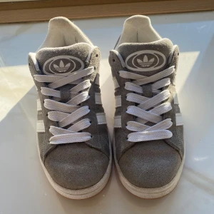 Grå Adidas Campus sneakers - Snygga grå Adidas Campus sneakers i mocka med vita detaljer och klassiska tre ränder på sidan. Skorna har en vit sula med gummifärg undertill och snörning framtill. Perfekta för en stilren look. Pris kan diskuteras 💖