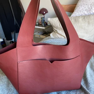 Carin Wester väska - Snygg röd axelväska i skinn med stilren design. Väskan har en praktisk öppen ficka på utsidan och en knappstängning inuti. Använd fåtal gånger💕