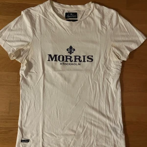 Vit t-shirt från Morris - Säljer en stilren vit t-shirt från Morris med tryck i svart på bröstet. T-shirten har en klassisk passform och är kortärmad, perfekt för en avslappnad stil.