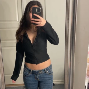 Svart ribbad kofta - Säljer en svart ribbad kofta med dragkedja framtill. Koftan har en croppad design och långa ärmar, perfekt för en stilren look. Passar bra till jeans eller kjol för en avslappnad stil.