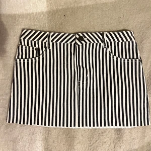 Randig kortkjol i jeansmaterial - Snygg kortkjol i svartvitt randigt mönster. Kjolen har en klassisk jeansdesign med fem fickor och bälteshällor. Perfekt för en trendig look!