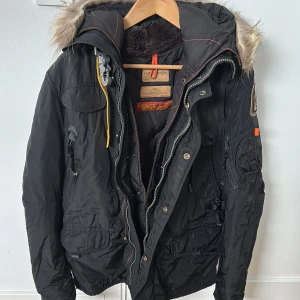 Svart vinterjacka från Parajumpers - Jag säljer en svart jacka från Parajumpers med pälsdetaljer på huvan och flera praktiska fickor. Jackan har dragkedja och knappar samt en unik gul remdetalj vid kragen. Perfekt för kalla dagar.  Skick 7/10