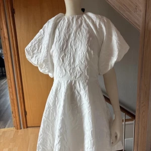 Vit klänning med puffärmar/ Studentklänning  - Säljer en elegant vit klänning med puffiga kortärmar och en knytning i ryggen. Klänningen har en texturerad yta som ger en unik look. Perfekt för speciella tillfällen!