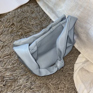 Ljusblå axelväska med knutdetalj - Säljer en stilren ljusblå axelväska i syntetmaterial. Väskan har en unik knutdetalj på handtaget och en rymlig insida. Perfekt för att bära dina dagliga nödvändigheter med stil.