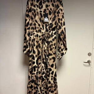 Leopardmönstrad långklänning från Black Colour - Snygg leopardmönstrad omlott långklänning från Black Colour i 100% polyester. Klänningen har långa ärmar och ett knytband i midjan för en smickrande passform. Perfekt för att sticka ut med stil! Ny 1299kr