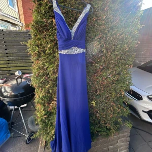 Blå långklänning med pärldetaljer - Elegant blå långklänning med v-ringad design och pärldetaljer vid midjan och halsen. Perfekt till bal eller fest. Storlek S/M