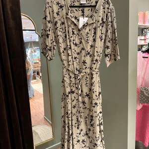 Beige blommig klänning från Humble - Snygg beige/grön klänning från Humble med svart blommönster. Klänningen har trekvartsärmar och ett knytband i midjan för en smickrande passform. Perfekt för en stilren look.