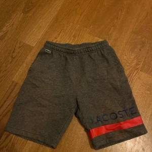 Grå shorts från Lacoste - Säljer ett par grå shorts från Lacoste med röd och blå detalj på benet. Hör av dig vid funderingar!