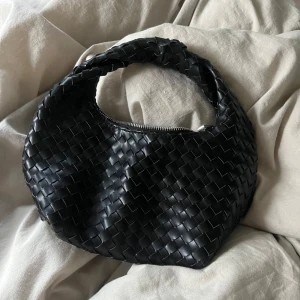 Svart flätad axelväska - Snygg svart axelväska med flätat mönster och dragkedja. Perfekt för att ge en stilren touch till din outfit. Väskan har en rundad form och ett bekvämt handtag.