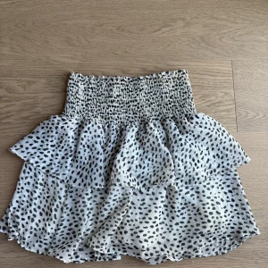 Vit och svart prickig kjol - Säljer en söt vit kjol med svarta prickar och volangdetaljer. Kjolen har en elastisk midja för bekväm passform. Perfekt för en somrig look!