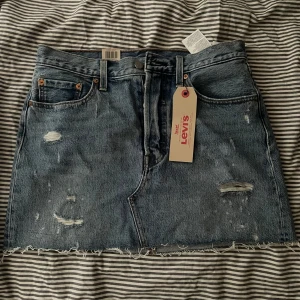 Jeanskjol från Levi's - Snygg jeanskjol från Levi's i en klassisk blå denim. Kjolen har en rå kant och slitna detaljer. Den har knappar framtill och femficksdesign. Aldrig använd.