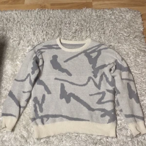Vit och grå stickad tröja - Säljer en vit och grå stickad tröja med ett abstrakt mönster. Tröjan har långa ärmar och ribbade muddar vid ärmslut och nederkant. Perfekt för kyliga dagar! Storlek M