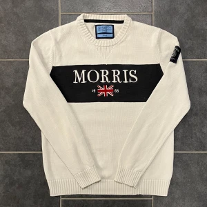 Vit tröja från Morris - Säljer en stilren vit tröja från Morris med svart panel och broderad logotyp samt Union Jack-flagga på bröstet. Tröjan har långa ärmar och ribbade muddar. Perfekt för en klassisk look.