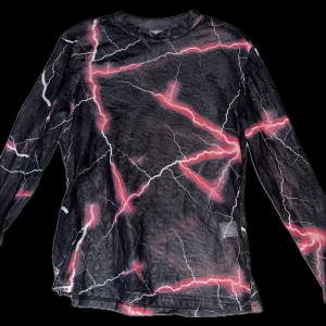 Svart och rosa långärmad tröja med blixtmönster - Cool långärmad tröja med ett unikt blixtmönster i rosa och vitt på en svart bakgrund. Perfekt för att sticka ut med en edgy stil. Den är även ganska genomskinlig så det är bra att man tänker på. Det är snyggt med ett svart linne eller tshirt under men funkar också utan. Använt en gång! 