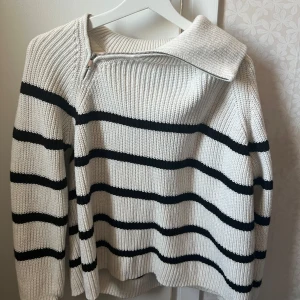 Vit och svart randig stickad tröja   - Säljer en stilren vit stickad tröja med svarta ränder. Tröjan har en unik dragkedja vid halsen och långa ärmar. Perfekt för kyligare dagar.