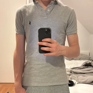 Grå pikétröja från Ralph Lauren - Säljer en klassisk grå pikétröja från Ralph Lauren med korta ärmar och en broderad logotyp på bröstet. Tröjan har en knappslå med två knappar och en stilren krage. Perfekt för en avslappnad men stilren look. Lite använd och inga defekter.