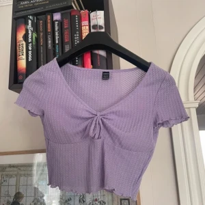 Lila kortärmad topp från Shein - Säljer en söt lila topp från Shein med korta ärmar och en knytning framtill. Toppen har en fin struktur och är perfekt för varmare dagar. Den är croppad och har en v-ringad design som ger en trendig look.