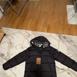 Svart dunjacka från Burberry - Säljer en stilren svart dunjacka från Burberry med avtagbara ärmar och klassiskt rutigt foder i huvan. Jackan har en dragkedja och knappar framtill samt en broderad logga på ärmen. Perfekt för kyliga dagar.