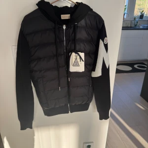 Svart cardigan/hybridjacka från Moncler - Säljer en snygg svart cardigan från Moncler med huva och dragkedja. Jackan har långa ärmar och en stor vit bokstav 'M' på ärmen. Perfekt för kyliga dagar. Knappt använd och etiketterna sitter kvar samt att påse finns också. NFC funkar också! Pris kan diskuteras! 